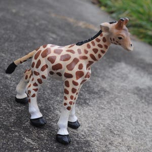 May include: A toy giraffe figurine with a tan and brown spotted pattern. The giraffe has white legs, black hooves, and a black tail. The figurine is standing on a gray surface.