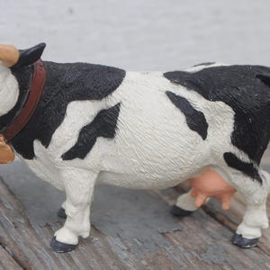 May include: A black and white cow figurine with a brown collar and bell. The cow has tan horns and a pink nose and udder. The figurine is standing on a wooden surface.