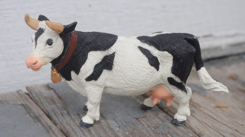 May include: A black and white cow figurine with a brown collar and bell. The cow has tan horns and a pink nose and udder. The figurine is standing on a wooden surface.