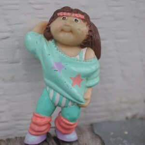 Könnte beinhalten: Eine Plastikfigur eines Cabbage Patch Kids, das ein türkisfarbenes und weißes Trainingsoutfit mit rosa Beinstulpen und einem rosa Stirnband trägt. Die Figur hat braunes Haar und einen rosa Stern auf ihrem Shirt.