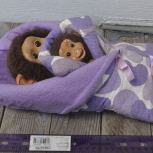 May include: Two plush monkey toys, one brown and one tan, are nestled together inside a purple and white patterned blanket. The blanket has a heart design and is tied with a pink ribbon.