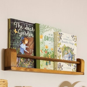 May include: A wooden bookshelf with three books. The books have colorful covers with titles like "The Secret Garden" and "We Are Garden." The shelf is made of light brown wood and is mounted on a white wall.