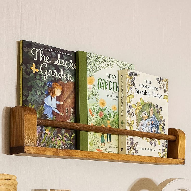 May include: A wooden bookshelf with three books. The books have colorful covers with titles like "The Secret Garden" and "We Are Garden." The shelf is made of light brown wood and is mounted on a white wall.