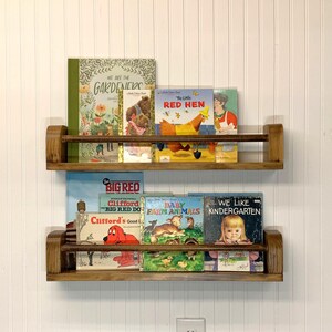 May include: Two wooden bookshelves mounted on a white wall, each holding a collection of children's books. The books feature colorful illustrations and titles like "The Little Red Hen" and "Clifford's Good Deeds".