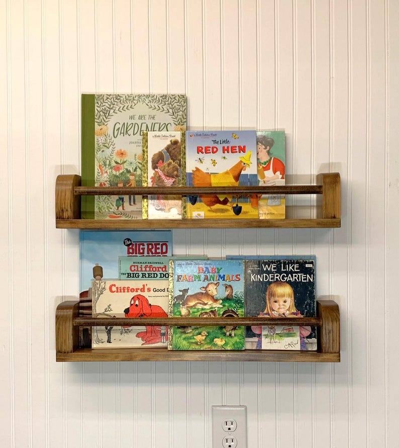 May include: Two wooden bookshelves mounted on a white wall, each holding a collection of children's books. The books feature colorful illustrations and titles like "The Little Red Hen" and "Clifford's Good Deeds".