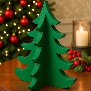 May include: A green, three-dimensional Christmas tree ornament. The tree is constructed from cut-out shapes and sits on a wooden table. A lit candle and a Christmas tree with red baubles are in the background.