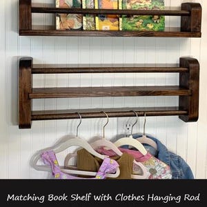 May include: Dark brown wooden wall-mounted bookshelf with two shelves. The top shelf holds vintage children's books. Below, a clothes hanging rod holds several children's garments. The text in the image says "Matching Book Shelf with Clothes Hanging Rod (Sold Separately)"