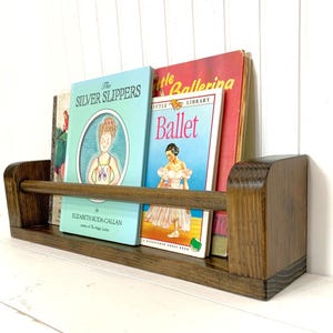 May include: A wooden bookshelf with a dark brown finish, holding several children's books. The books have colorful covers, including titles like "The Silver Slippers" and "Ballet." The shelf is mounted on a white wall.