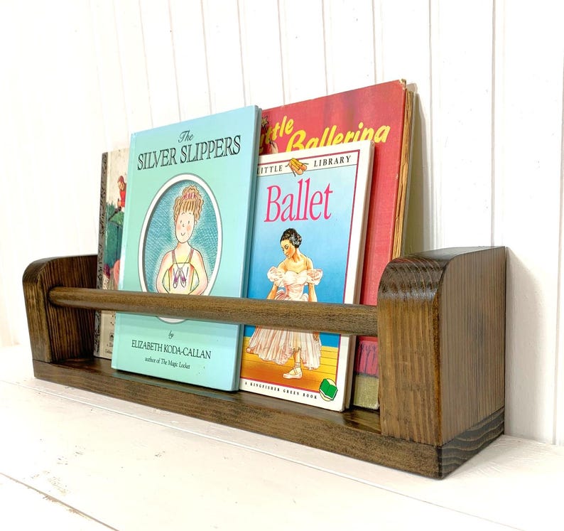 May include: A wooden bookshelf with a dark brown finish, holding several children's books. The books have colorful covers, including titles like "The Silver Slippers" and "Ballet." The shelf is mounted on a white wall.