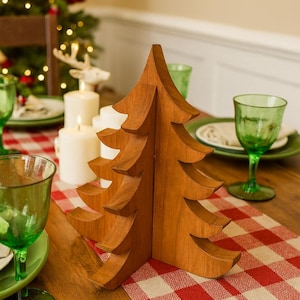 May include: A wooden Christmas tree ornament sits on a red and white checked table runner. The tree is made of light brown wood and has a layered design. Green glassware and candles are also on the table.