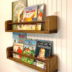 May include: Two wooden bookshelves mounted on a white wall, each holding several children's books. The shelves are stained a warm brown and have a lip to keep books in place. The books have colorful covers.