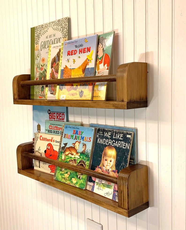 May include: Two wooden bookshelves mounted on a white wall, each holding several children's books. The shelves are stained a warm brown and have a lip to keep books in place. The books have colorful covers.