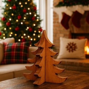 May include: A wooden Christmas tree ornament with a layered design, placed on a dark wooden table. The tree is in front of a decorated Christmas tree and a fireplace with stockings.