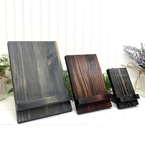 May include: Three wooden display stands in varying sizes and shades of gray and brown. Each stand has a rectangular base and a slightly angled back, with a small ledge at the bottom to hold items. The wood grain is visible.