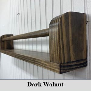 May include: A dark walnut-stained wooden shelf with a rod. The shelf has a rectangular base and rounded end supports. The rod is positioned above the shelf, and the text "Dark Walnut" is at the bottom.