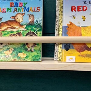 May include: A light-colored wooden bookshelf with a rounded front edge and a wooden dowel to keep books in place. The shelf holds three children's books with colorful covers, including "I Love You, Grandma!" and "The Little Red Hen."
