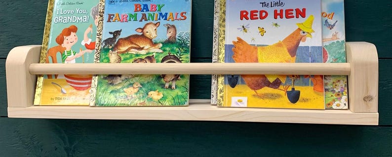 May include: A light-colored wooden bookshelf with a rounded front edge and a wooden dowel to keep books in place. The shelf holds three children's books with colorful covers, including "I Love You, Grandma!" and "The Little Red Hen."