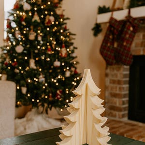May include: A wooden Christmas tree ornament stands on a green table. A decorated Christmas tree and fireplace are in the background. The wooden tree is light-coloured and has a layered design. The room is decorated for the holidays.