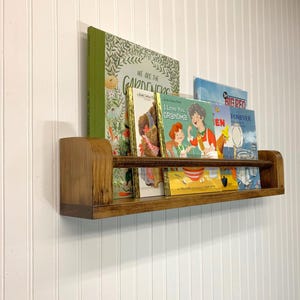 May include: A wooden bookshelf with a curved front edge, holding several children's books. The books have colorful covers, including titles like "We Are the Gardeners" and "I Love You, Grandma!". The shelf is mounted on a white paneled wall.