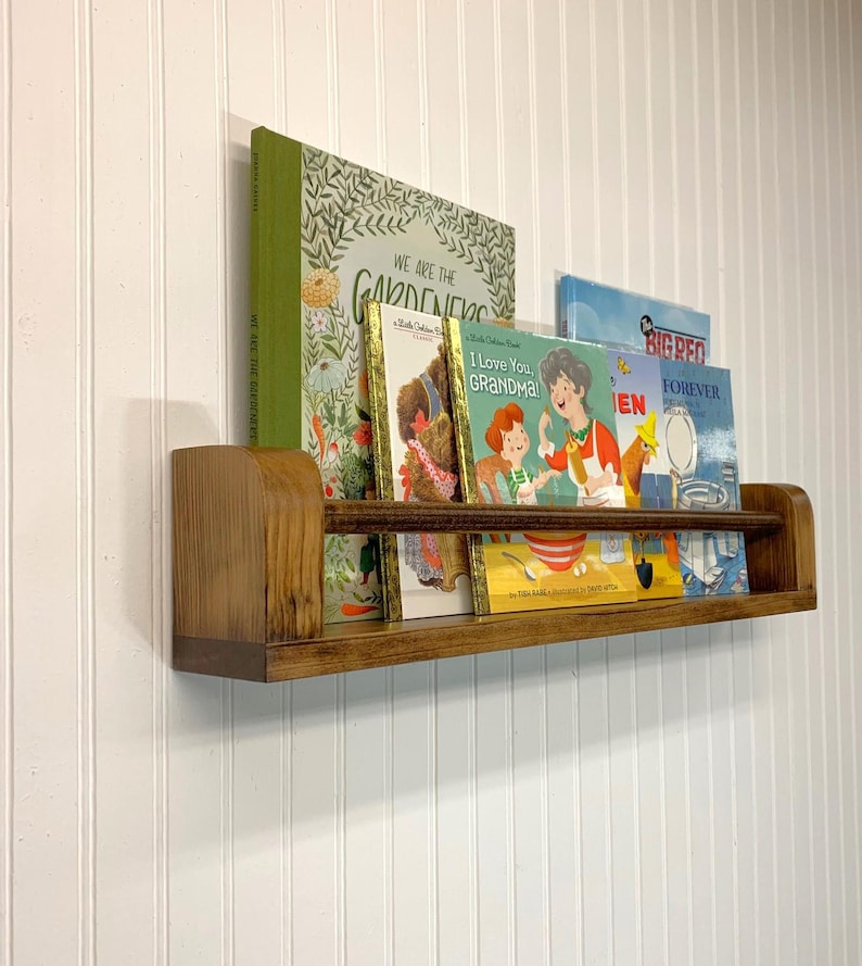 May include: A wooden bookshelf with a curved front edge, holding several children's books. The books have colorful covers, including titles like "We Are the Gardeners" and "I Love You, Grandma!". The shelf is mounted on a white paneled wall.