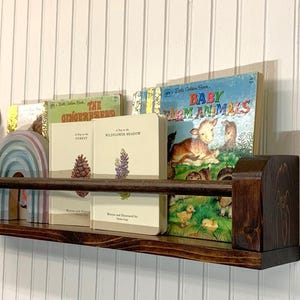 May include: A dark stained wooden bookshelf with a selection of children's books, a rainbow-shaped wooden toy, and a purple bow. The books include titles like "Baby Farm Animals" and "The Gingerbread Man."