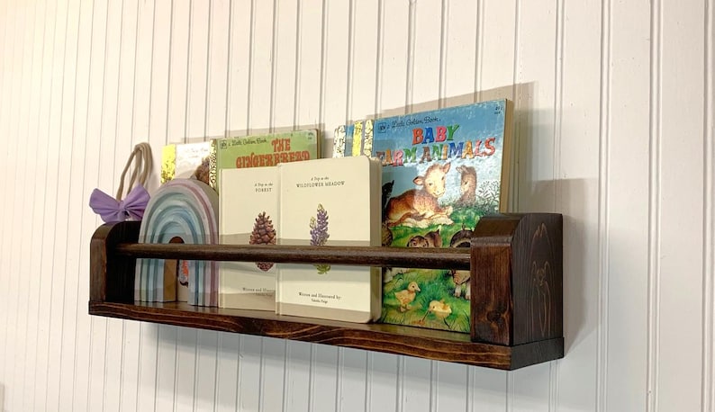 May include: A dark stained wooden bookshelf with a selection of children's books, a rainbow-shaped wooden toy, and a purple bow. The books include titles like "Baby Farm Animals" and "The Gingerbread Man."