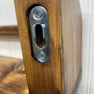 May include: Close-up of a stained wooden shelf with a metal bracket for wall mounting. The wood has a rich, dark brown finish. The bracket is silver and secured with screws, designed for easy installation.