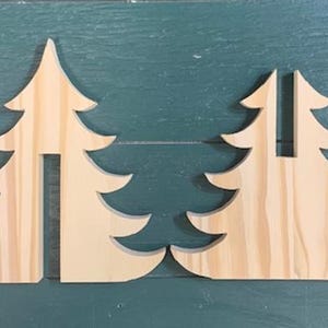 May include: Two unfinished wooden Christmas tree cutouts. Each tree has a cutout in the centre. The trees are light brown and sit on a teal-coloured wooden surface. The trees are designed with layered branches and a pointed top.