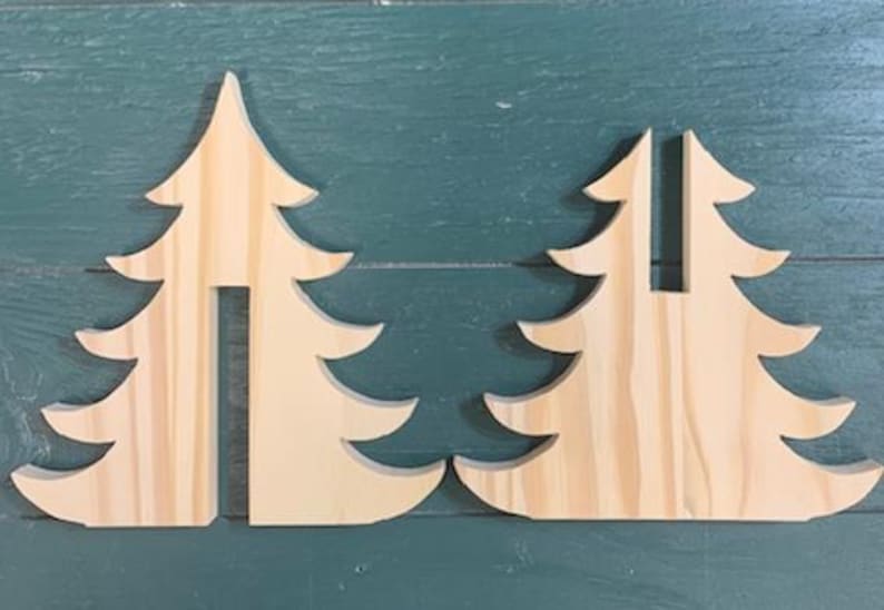 May include: Two unfinished wooden Christmas tree cutouts. Each tree has a cutout in the centre. The trees are light brown and sit on a teal-coloured wooden surface. The trees are designed with layered branches and a pointed top.