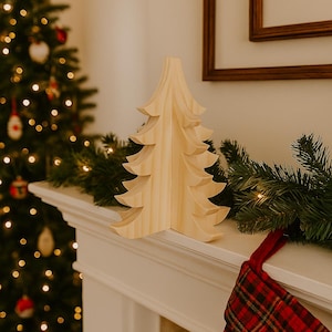 May include: A wooden Christmas tree ornament sits on a white mantelpiece, with a decorated Christmas tree and a red plaid stocking in the background. The wooden tree is light-coloured and has a layered design.