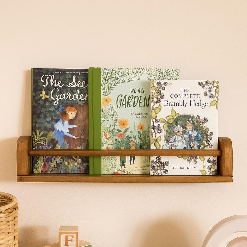 May include: A wooden bookshelf displays three hardcover books. The books are titled "The Secret Garden", "We Are Garden", and "The Complete Brambly Hedge". The bookshelf is made of light brown wood and is mounted on a beige wall.