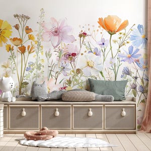 May include: A floral mural with watercolor-style flowers in shades of yellow, orange, pink, and blue adorns a wall. Below, a storage unit with woven doors and round knobs holds decorative pillows and a white bear lamp. A pink curtain hangs on the right.