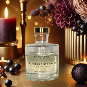 May include: A clear glass bottle with a gold lid and label that reads "LIWUMI HANDCRAFTED REED DIFFUSER MIDNIGHT STARDUST STRAWBERRY RED CURRANT BLACK TEA MADE BY HAND IN CONNECTICUT WITH LOVE 6.7 FL OZ". The bottle is sitting on a gold surface with a candle and other decorative items.