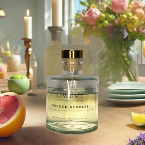 May include: A clear glass reed diffuser with a gold cap and the text "Brunch Bubbles" on the label. The diffuser is surrounded by a halved grapefruit, green apples, and a glass of orange juice. A bouquet of flowers is in the background.