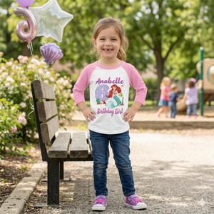 May include: A young girl wearing a white and pink raglan shirt with the text "Anabelle 6 Birthday Girl" and a graphic of a mermaid. She is wearing blue jeans and pink sneakers. Balloons are in the background.