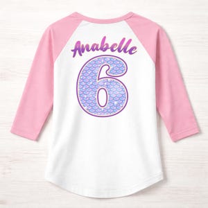 May include: A white and pink raglan shirt with the name "Anabelle" and the number "6" on the back. The number is designed with a mermaid scale pattern in shades of purple and blue. The sleeves are pink.