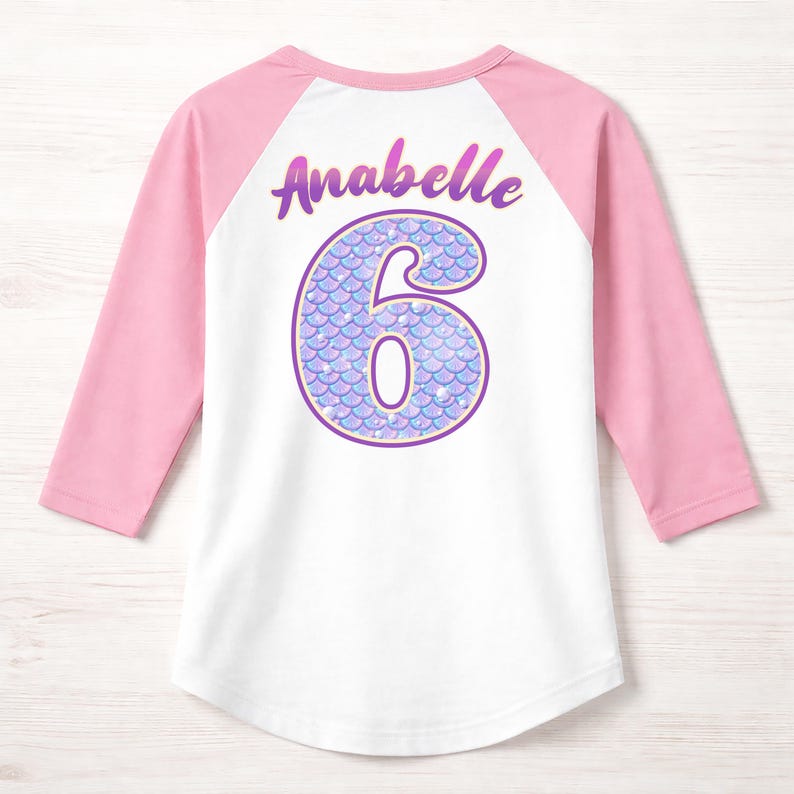 May include: A white and pink raglan shirt with the name "Anabelle" and the number "6" on the back. The number is designed with a mermaid scale pattern in shades of purple and blue. The sleeves are pink.