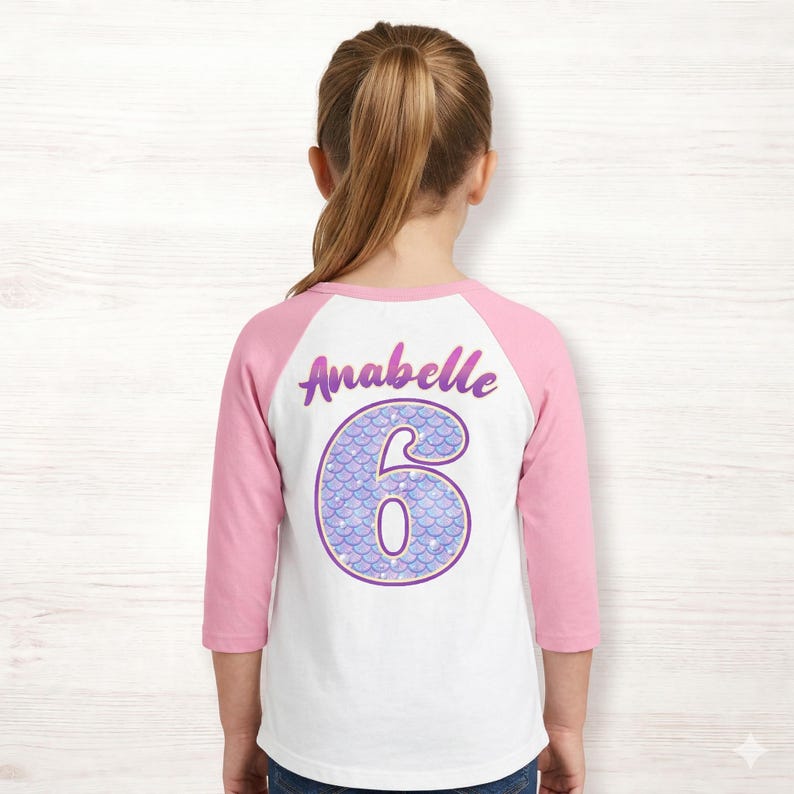 May include: White and pink raglan style shirt with the name "Anabelle" and the number "6" in a mermaid scale design. The number is purple with a light blue and white scale pattern.