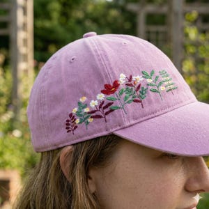 May include: A lavender baseball cap with embroidered floral design. The embroidery features red, white, and yellow flowers with green leaves. The cap has a curved brim and a rounded crown.