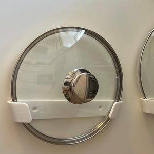 Könnte beinhalten: Zwei Deckel aus klarem Glas mit Edelstahlrändern und -griffen sind an einer weißen Wand montiert. Die Deckel werden von weißen Kunststoffhaltern gehalten. Ein Deckel hat einen großen, runden Griff, der andere einen schlaufenförmigen Griff.