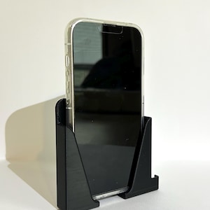 Könnte beinhalten: Ein schwarzer Telefonständer mit einer durchsichtigen Handyhülle darin. Der Ständer ist aus Kunststoff und hat eine strukturierte Oberfläche.