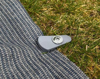 May include: A close-up of a grey fabric with a silver metal clip. The clip has a round, silver button with a screw in the centre.