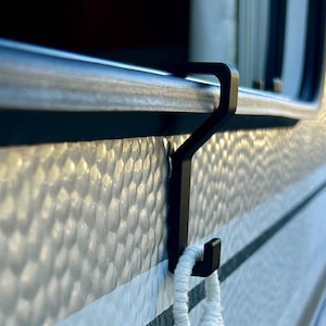 Könnte beinhalten: Ein schwarzer Metallhaken hängt an einer strukturierten Wand und hält eine weiße Makramee-Schlaufe. Der Haken ist so konzipiert, dass er über einer horizontalen Stange hängt, wahrscheinlich für ein Handtuch oder andere kleine Gegenstände.