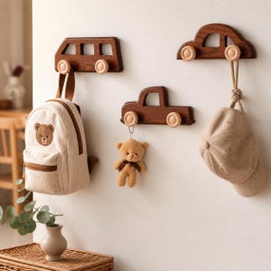 Könnte beinhalten: Drei Wandhaken in Form von Autos aus dunkelbraunem Holz mit hellen Holzrädern. Ein beiger Rucksack mit Bärenmuster, ein Teddybär und eine beige Kappe hängen daran, was eine charmante Aufbewahrungslösung darstellt.