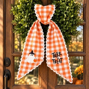 May include: An orange and white gingham fabric bow with scalloped edges. The left tail features a white ghost wearing a black witch hat and holding an orange pumpkin. The right tail displays the black text Trick or Treat in a stylized font.