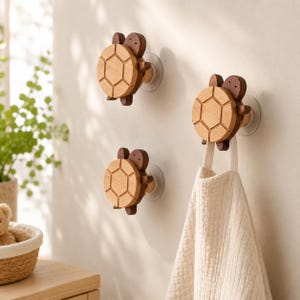 May include: Three wooden turtle-shaped wall hooks with suction cups. The turtle shells are light brown with a geometric design, and the heads and limbs are a darker brown. One hook holds a cream-colored towel.