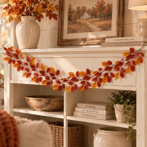 Puede incluir: Una guirnalda decorativa con hojas de otoño artificiales en tonos rojos, naranjas y amarillos, colocada sobre una repisa de chimenea de madera blanca. La guirnalda consta de tallos marrones finos con numerosas hojas pequeñas y ovaladas.
