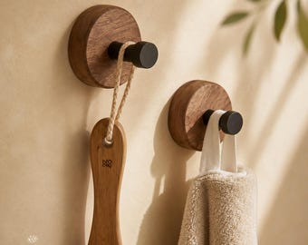 May include: Two wooden wall hooks with black metal pegs. One hook holds a wooden brush with a rope loop, the other holds a beige towel. The hooks are mounted on a light beige wall. The text "Sweet Timber Decor" is in the lower left corner.