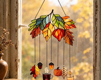 Op de afbeelding: Een glas-in-lood zonnevanger met een cluster van veelkleurige herfstbladeren aan de bovenkant, met hangende bedels waaronder extra bladeren, eikels, een dampende mok, een pompoen en een kaars, allemaal opgehangen aan dunne metalen kettinkjes.