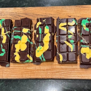 May include: Five dark chocolate bars with colorful yellow and green drizzle are arranged on a wooden cutting board. The bars are rectangular with individual squares, and the drizzle adds a decorative touch. The wooden board has a logo in the bottom left corner.